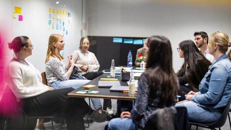 Sieben Personen sitzen in einem Tagungsraum um einen Tisch und unterhalten sich. An der Wand: Bunte Post-its.
