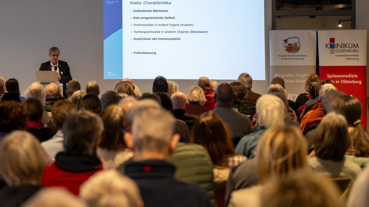 Volle Sitzreihen, im Hintergrund ist ein Referent neben einer Leinwand zu erkennen.