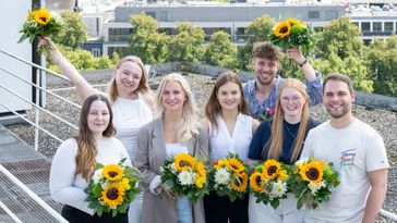 Fünf Frauen und zwei Männer mit Sonnenblumen-Sträußen lächeln in die Kamera.
