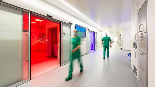 Ein langer Flur, zwei Personen in grüner OP-Kleidung bewegen sich darin. Eine Schiebetür steht offen, der Raumdahinter ist rot beleuchtet. Aus einem weiter hinten liegenden Raum scheint es bläulich-violett. 