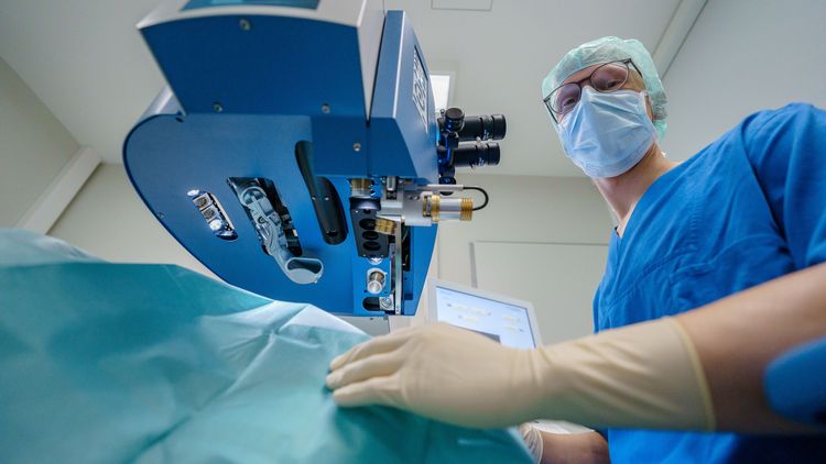 Ein Arzt in Kasak mit Haube und Mundschutz steht neben einem Augenlaser. Das Bild ist aus Patientenperspektive, also von unten, aufgenommen.