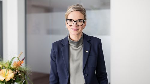 Porträtbild von Nadine Krefeld mit Hochsteckfrisur, schwarzer Brille, blauem Blazer und grauem Rollkagenpullover. Sie lächelt freundlich in die Kamera.