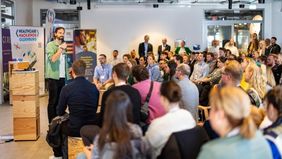 Mehrere Personen in einer Art Vortragssituation. Vorn steht ein Sprecher mit Mikro und grünem Hand, neben ihm ein Roll-up mit der Aufschrift "Healthcare Hackathon".