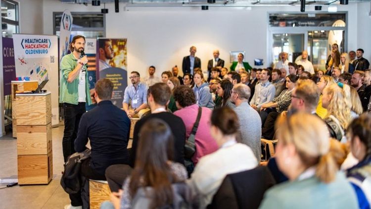 Mehrere Personen in einer Art Vortragssituation. Vorn steht ein Sprecher mit Mikro und grünem Hand, neben ihm ein Roll-up mit der Aufschrift "Healthcare Hackathon".