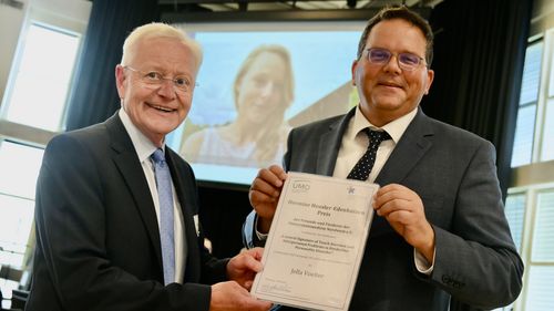 Norbert Wahn überreicht die Urkunde an René Hurlemann. Im Hintergrund ist Jelle Voelter als Video auf einer Leinwand zu sehen. 
