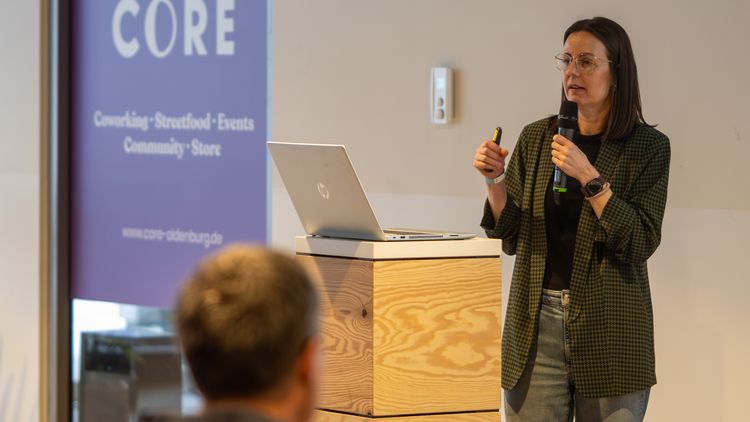 Eine Referentin steht an einem Stehpult, auf dem ein Laptop steht. Im Vordergrund ist ein Hinterkopf zu erkennen.