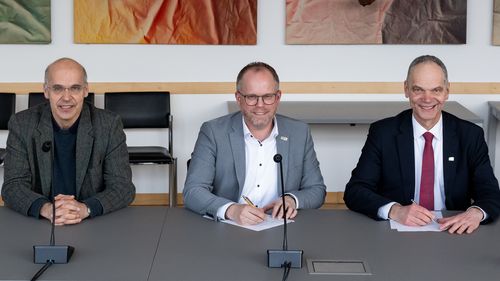 Nothwang, Leenderts und Bruder sitzen im Senatssitzungssaal. Vor Leenderts und Bruder liegen Dokumente, die sie unterschreiben. Alle schauen in die Kamera.