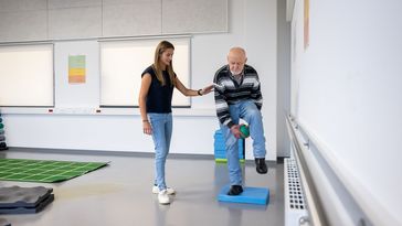 Eine Frau assistiert einem älteren mann, der auf einer weichen Matte steht, ein Bein anhebt und darunter einen Ball hindurchführt.