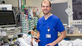 Johannes Grone in einem Intensivstationszimmer. Hinter ihm im Bett liegt eine Simulationspuppe, die von vielen Geräten umgeben ist.