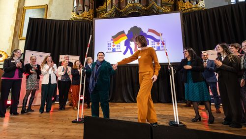 Zwei Frauen durchschreiten auf einer Bühne nach oben geklappte Schlagbäume. Im Hintergrund stehen Menschen und applaudieren.