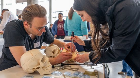Ein Mann und eine junge Frau sind über einen Schädel gebeugt, an dem Metallteile zu sehen sind, die sie mit einem Schraubenzieher bearbeiten.