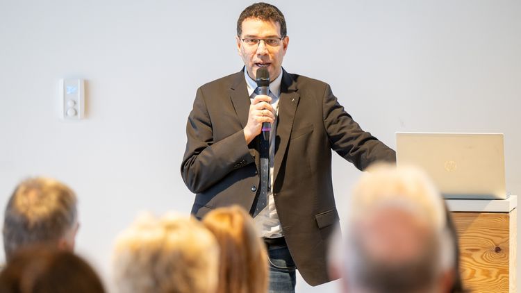 Cyrus Khandanpour im Sakko mit Mikrofon, in das er spricht. Im Vordergrund: Hinterköpfe.