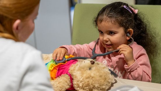 Vor einem kleinen Mädchen liegt ein Teddy mit buntem Kleid. Das Mädchen untersucht ihn mit einem Stethoskop.