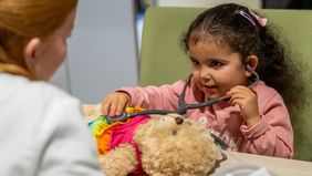 Vor einem kleinen Mädchen liegt ein Teddy mit buntem Kleid. Das Mädchen untersucht ihn mit einem Stethoskop.