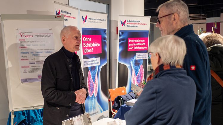 Ein Mann mit grauem Haar steht hinter dem Infostand und unterhält sich mit einem ebenfalls grauhaarigen Paar auf der anderen Seite des Tisches.