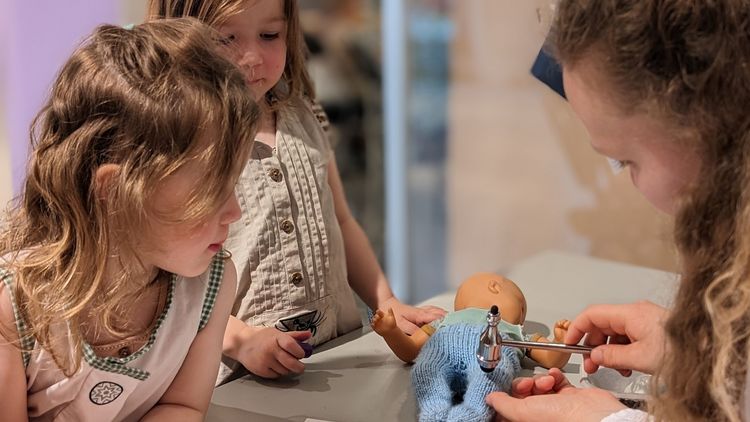 Das Bild zeigt zwei kleine Mädchen, die einer Medizinstudentin dabei zuschauen, wie sie bei einer Puppe die Reflexe untersucht.