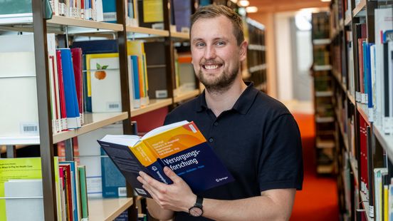 Jannik Ohmes in der Bibliothek der Uni Oldenburg. Er hält ein Buch mit dem Titel "Versorgungsforschung" in der Hand.