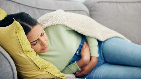 Frau liegt auf Sofa und krümmt sich vor Schmerzen. Sie hält sich den Bauch.