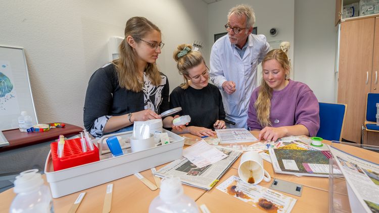 Die drei Studentinnen sitzen am Tisch, der voll ist mit medizinischen Materialien wie Mundspatel, Spritzen und Infusionsbehältern. Hinter ihnen steht ein Mann im Kittel.