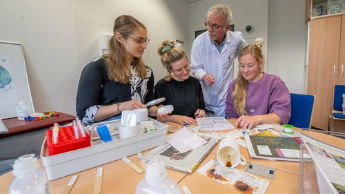 Die drei Studentinnen sitzen am Tisch, der voll ist mit medizinischen Materialien wie Mundspatel, Spritzen und Infusionsbehältern. Hinter ihnen steht ein Mann im Kittel.
