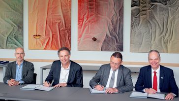 Die vier genannen Personen tragen Anzüge und sitzen nebeneinander an einem Tisch. Jouke de Vries signiert. Im Hintergrund hängen vier bunte Bilder.