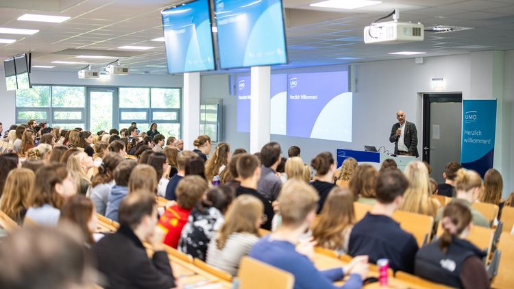 Ein gut gefüllter Hörsaal. Die Studierenden sind von hinten zu sehen. Vorn steht ein Mann mit Mikrofon. Neben ihm ein Roll-up mit der Aufschrift: UMO - Herzlichen willkommen. 