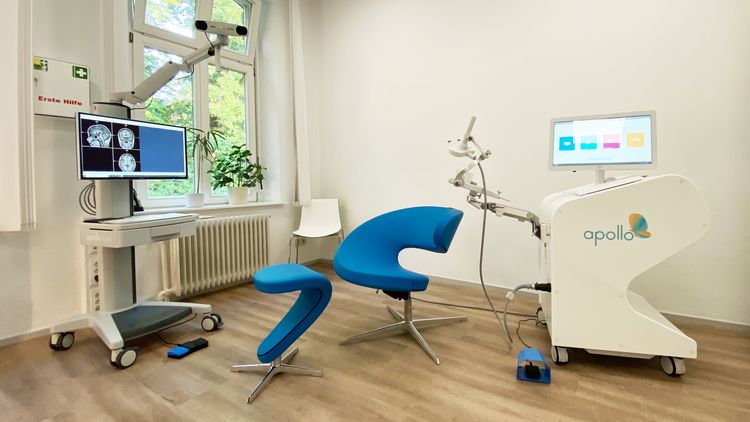 Links ein Monitor mit Gehirnscans, in der Bitte ein Stuhl mit Fußhocker, rechts das TMS-Gerät: Auf einem Wagen steht ein Monitor, am Wagen ist außerdem ein Arm befestigt, der die hutähnliche Apparatur trägt, die während der Therapie den Kopf umgibt.
