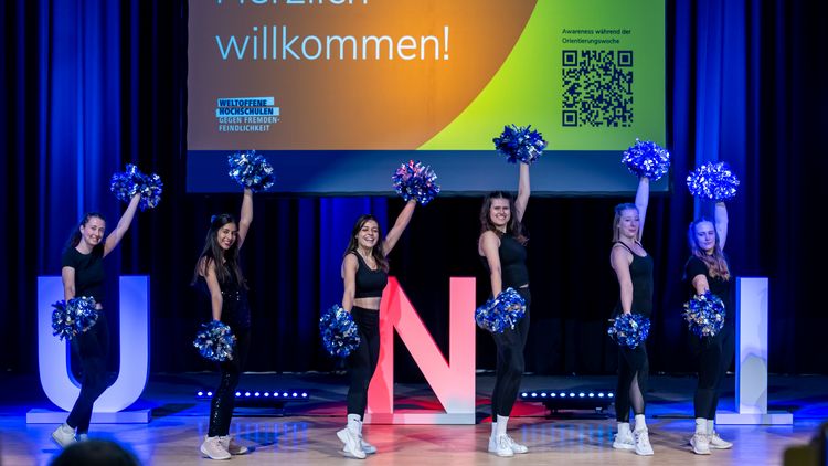 Cheerleader strecken Pompons nach oben und unten. Hinter ihnen steht auf einer Leinwand: Willkommen.