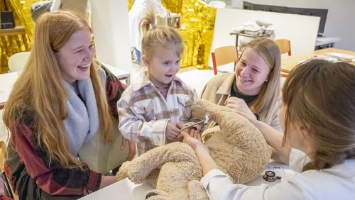 Ein kleines Mädchen hält einen Reflexhammer. Vor ihr liegt ein großer Plüschhund. Rechts und  links neben ihr sitzen zwei lächelnde Frauen. Ihr gegenüber sitzt eine Frau im Kittel.