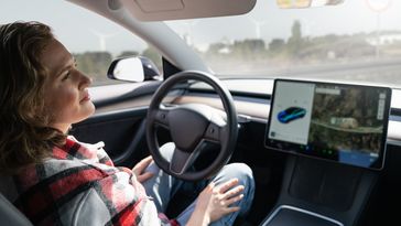 Frau auf dem Fahrersitz eines Autos. Sie berührt das Lenkrad nicht. Neben dem Lenkrad ist ein großes Display, das das Fahrzeug von außen und die Umgebung zeigt.