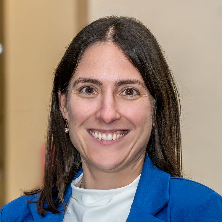 Porträt Jasmina Sterz. Sie hat schulterlange dunkle Haare, trägt ein weißes Shirt und einen blauen Blazer.