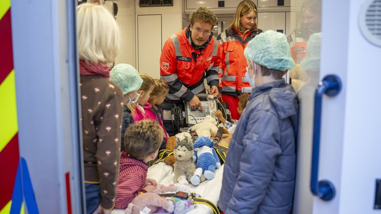 Kinder stehen im Rettungswagen um die Trage herum, auf der ihre Kuscheltiere festgeschnallt sind. Ein Rettungssanitäter erklärt etwas.