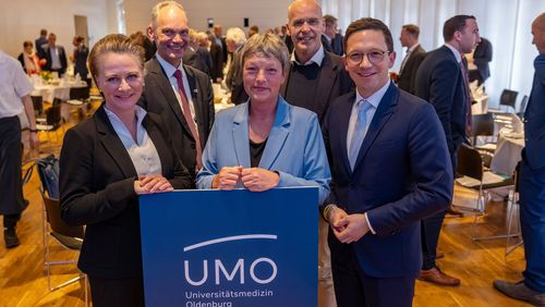 Die genannten Personen stehen hinter einem Schild, auf dem das UMO-Logo zu sehen ist. Im Hintergrund ist der Saal des Alten Rathauses zu sehen, in dem sich mehrere Personen miteinander unterhalten.