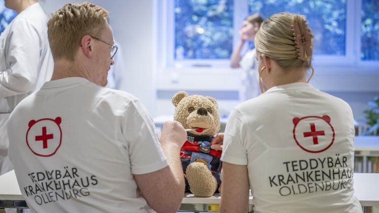 Zwei Medizinstudierende von hinten. Auf ihren T-Shirts steht "Teddybärkrankenhaus Oldenburg". Zwischen ihnen ist ein Plüschteddy zu erkennen, den sie gerade untersuchen.