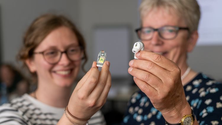 Eine junge und eine etwas ältere Frau halten jeweils ein kleines  Gerät in die Kamera, das wie ein In-ear-Kopfhörer aussieht.